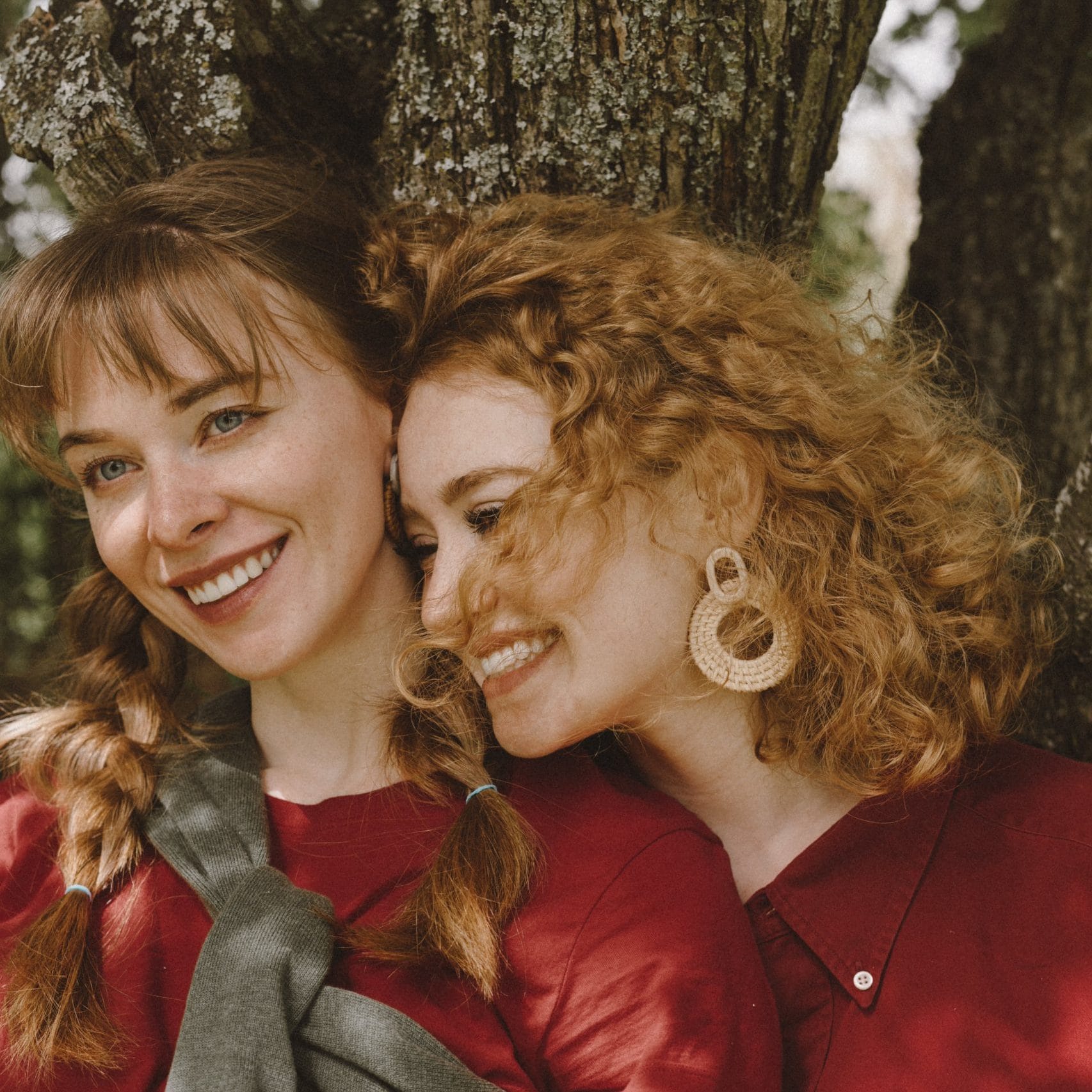 Illustration: Two women in red dresses standing close and laughing in front of a large tree trunk.